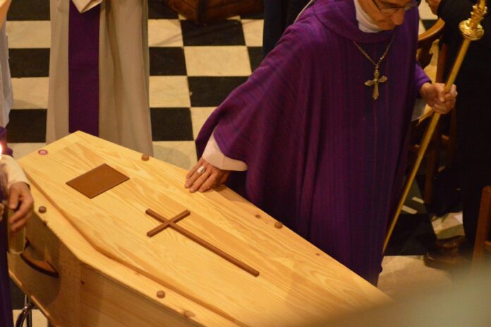 Ein Geistlicher legt seine Hand auf einen Holzsarg mit einem Kreuz.