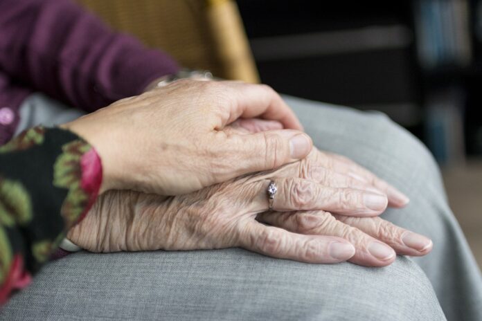 Eine Hand einer jüngeren Person liegt auf den Händen einer älteren Person auf dem Knie.