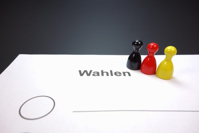 Wahlschein mit drei kleinen Spielfiguren in den Farben Schwarz, Rot und Gelb.